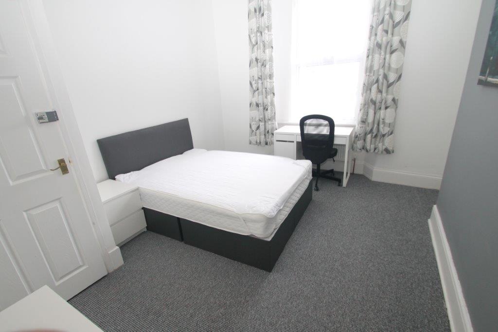 Second student bedroom with double bed, white desk with black office chair in front of window, bedside table and white chest of drawers